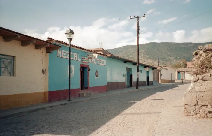 A row of connected single story colorful buildings with only one sign on it saying "Mezcal." The road is dirt and stone with one old lamp post and one electrical post.